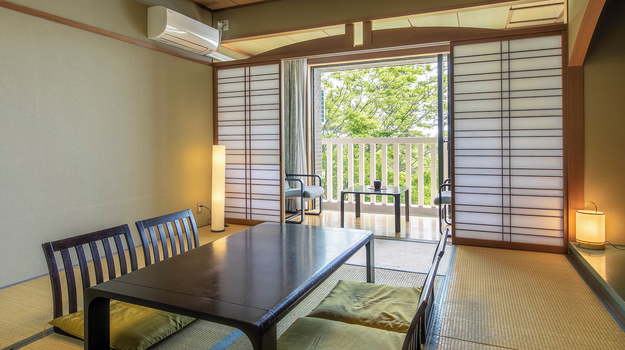 A traditional Japanese room with tatami flooring, a low table, and a sliding door leading to a balcony with a view of greenery.