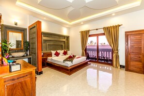 A spacious bedroom with a large bed, a wooden dresser, a vanity with a mirror, and a view through a glass door.
