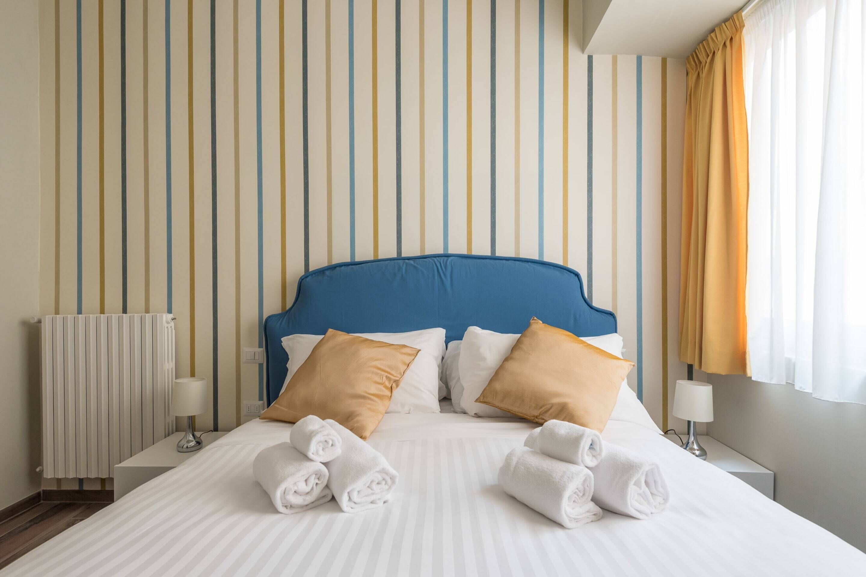 Une chambre d’hôtel avec un mur à rayures, une tête de lit bleue, du linge de lit blanc et deux lampes de chevet.