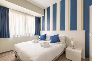 A hotel room with a large bed, blue pillows, white linens, a nightstand with a lamp, and a window with curtains. - Signoria Conte's Suite (Florence)