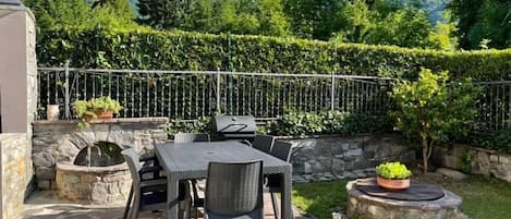 An outdoor patio with stone flooring, a table and chairs, a grill, and a view of mountains and greenery.