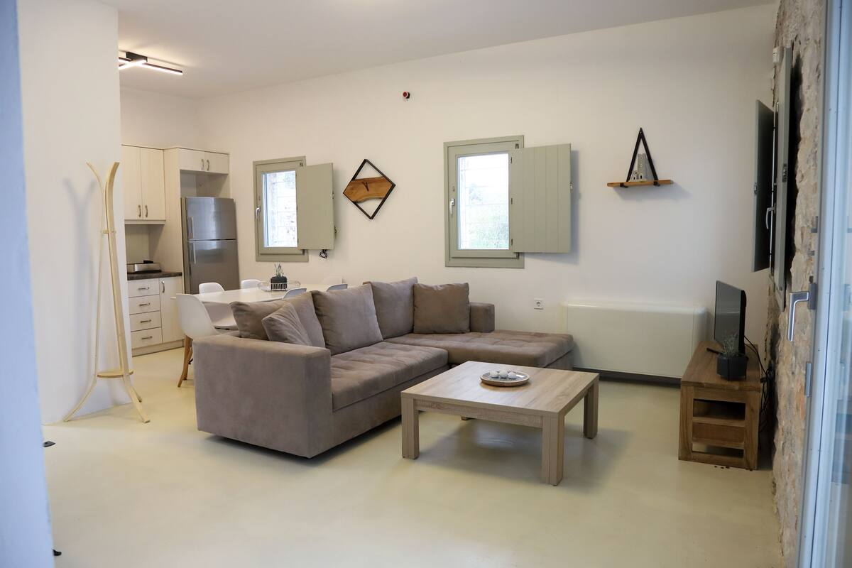 Un salon moderne avec un canapé d’angle, une table basse en bois et un coin cuisine équipé de placards et d’un réfrigérateur.
