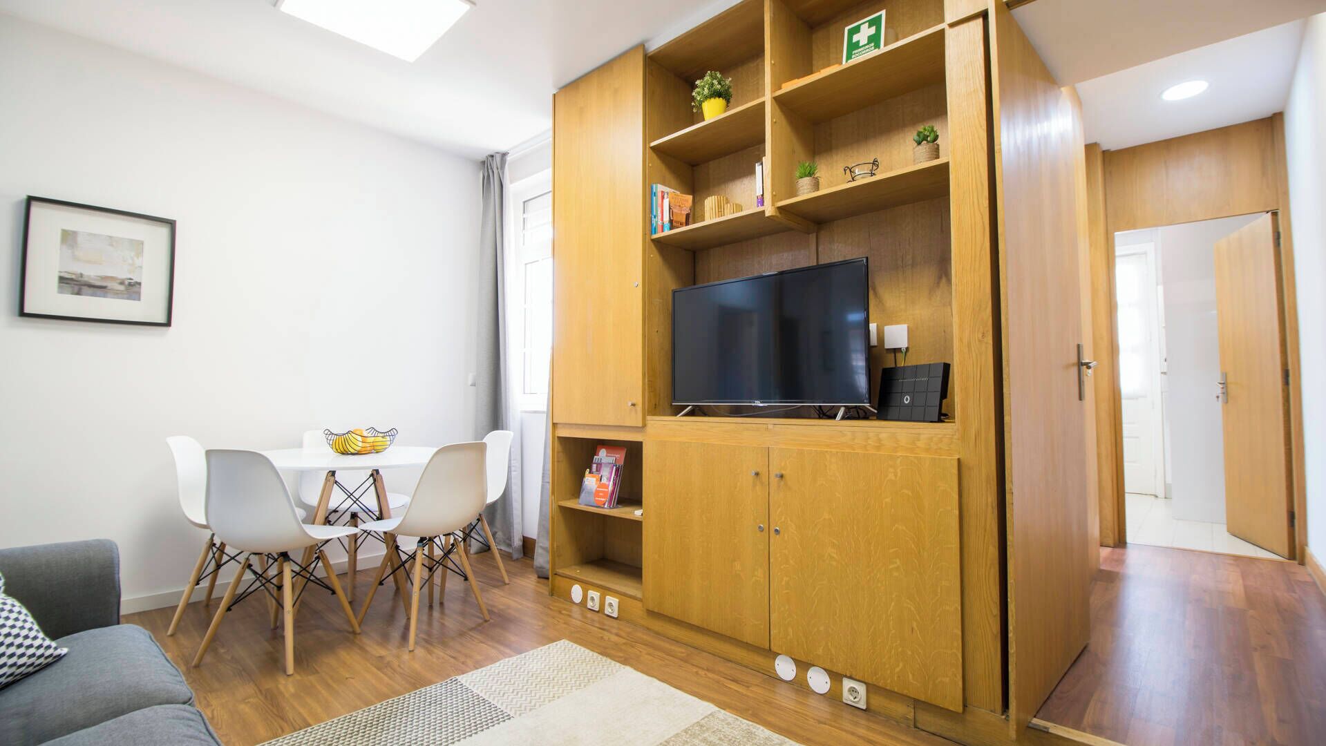 Ein modernes Wohnzimmer mit einer Holzeinrichtungsanlage, einem Flachbildfernseher, einer Essecke mit weißem Tisch und Stühlen sowie einem grauen Sofa.