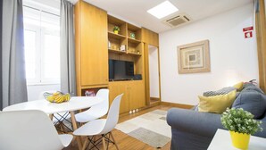 Un salon moderne avec un canapé, une table à manger et des chaises, un téléviseur posé sur un meuble en bois, et une petite plante sur le canapé.