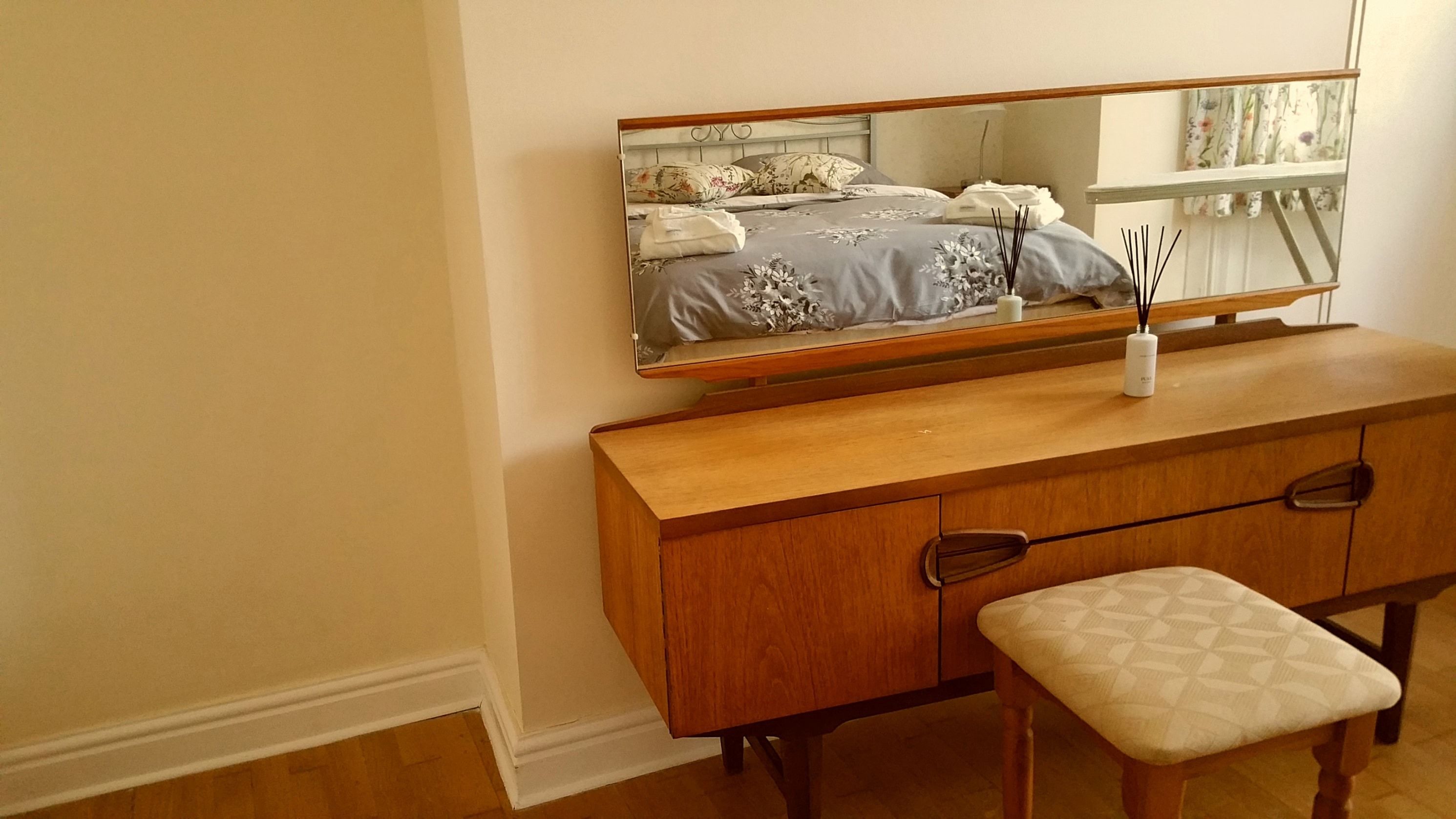 A wooden dresser with a mirror above it, a small bench, and a bed with floral bedding in the reflection.