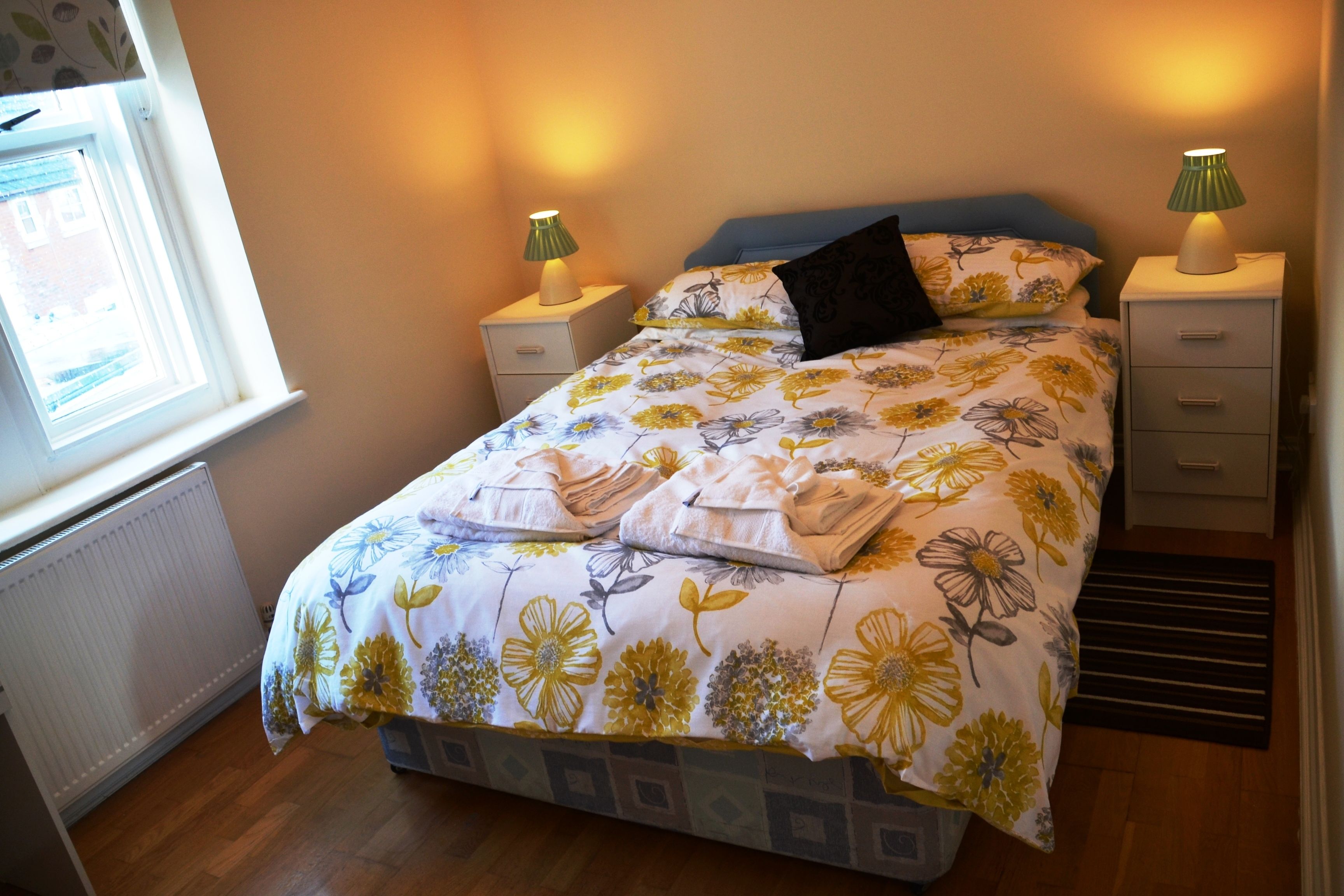 A bedroom with a floral bedspread, two bedside lamps, a window, and a radiator.