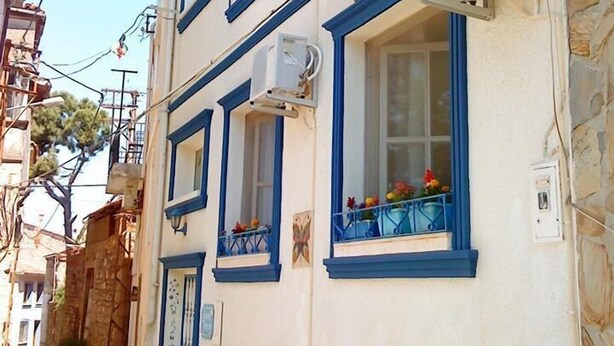A two-story building with blue-framed windows and balconies, stone walls, and a narrow street. - Kelebek Ayvalık (Ayvalik)