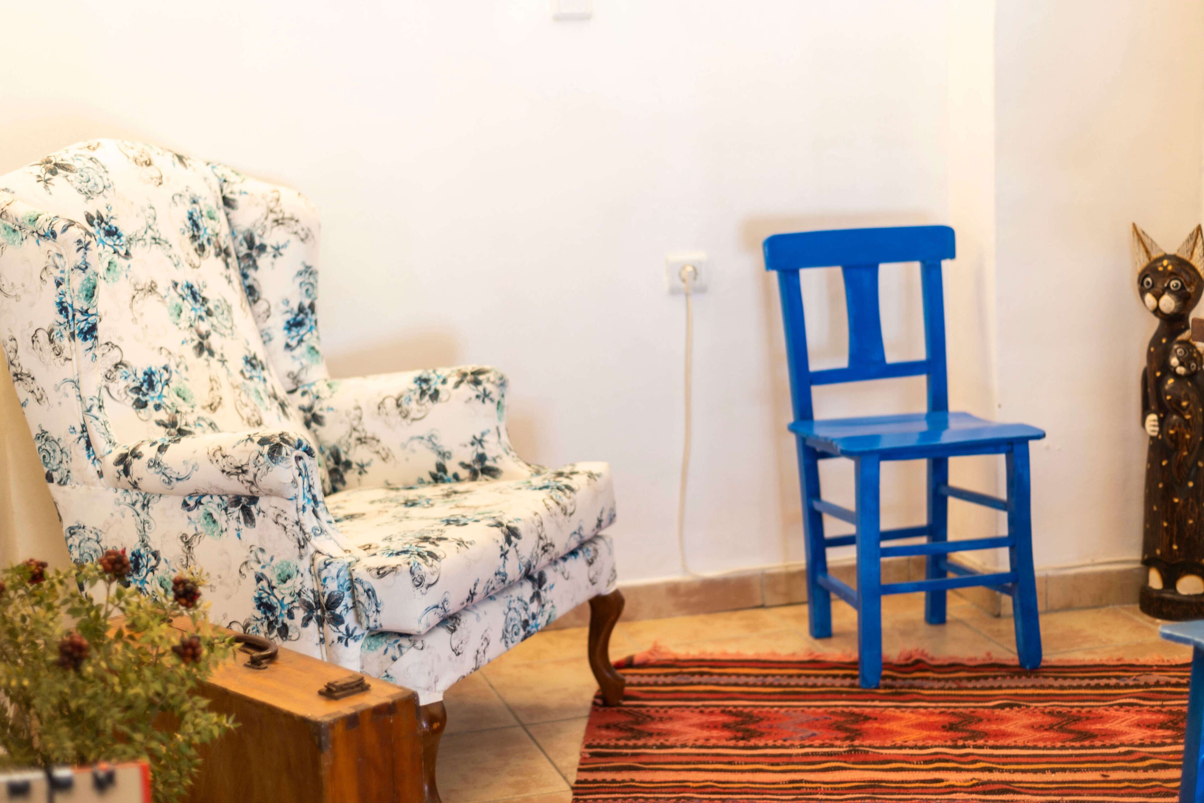 Ein Zimmer mit einem blumigen Sessel, einem blauen Stuhl, einem Holztisch und einem roten Teppich mit Muster.