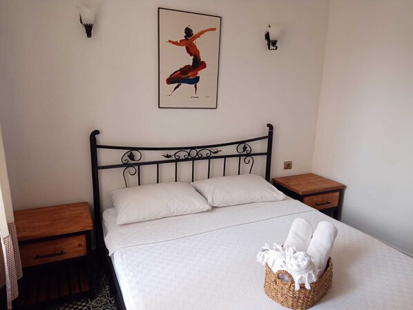 A bedroom with a bed, two nightstands, a wooden table, a basket with towels, and a wall-mounted painting.