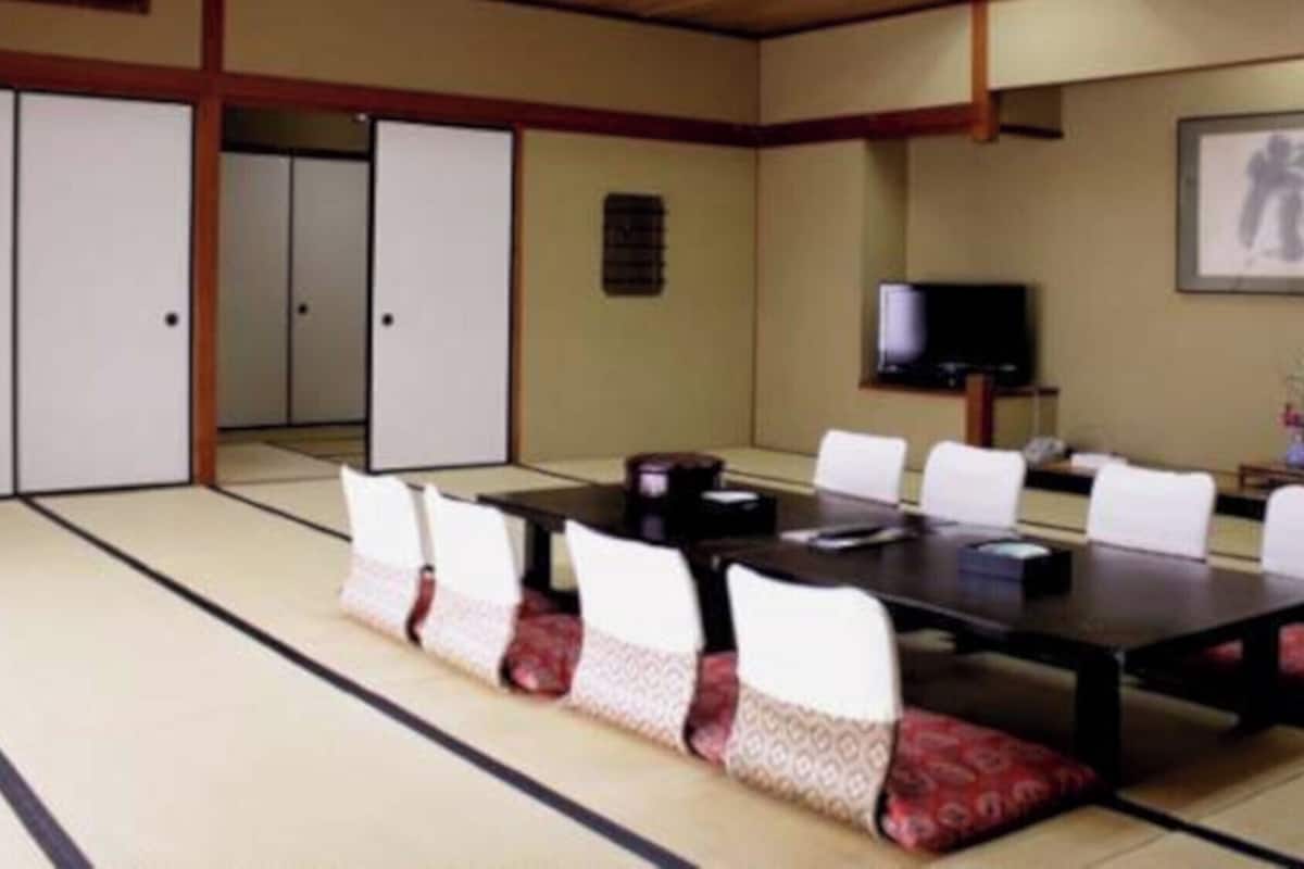 Ein traditioneller japanischer Raum mit Tatami-Böden, einem niedrigen Tisch mit Stühlen, einem Fernseher und einem gerahmten Kunstwerk an der Wand.
