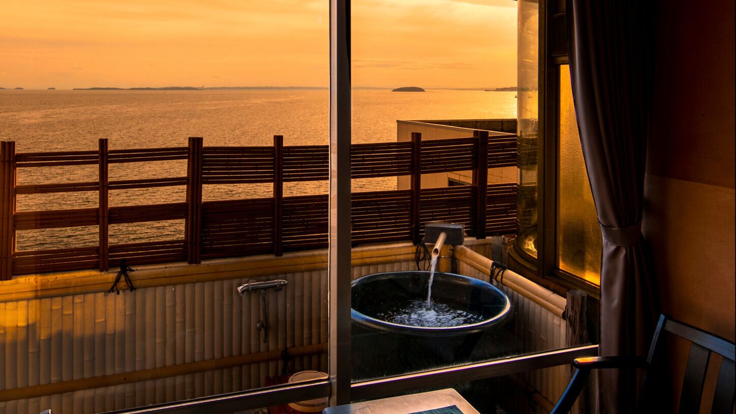 Une pièce avec vue sur la mer au coucher du soleil, une fenêtre avec un garde-corps en bois et un lavabo avec de l’eau courante.