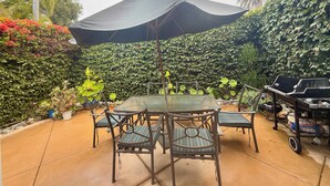 A patio with a table and chairs under a large umbrella, surrounded by lush greenery and a barbecue grill.
