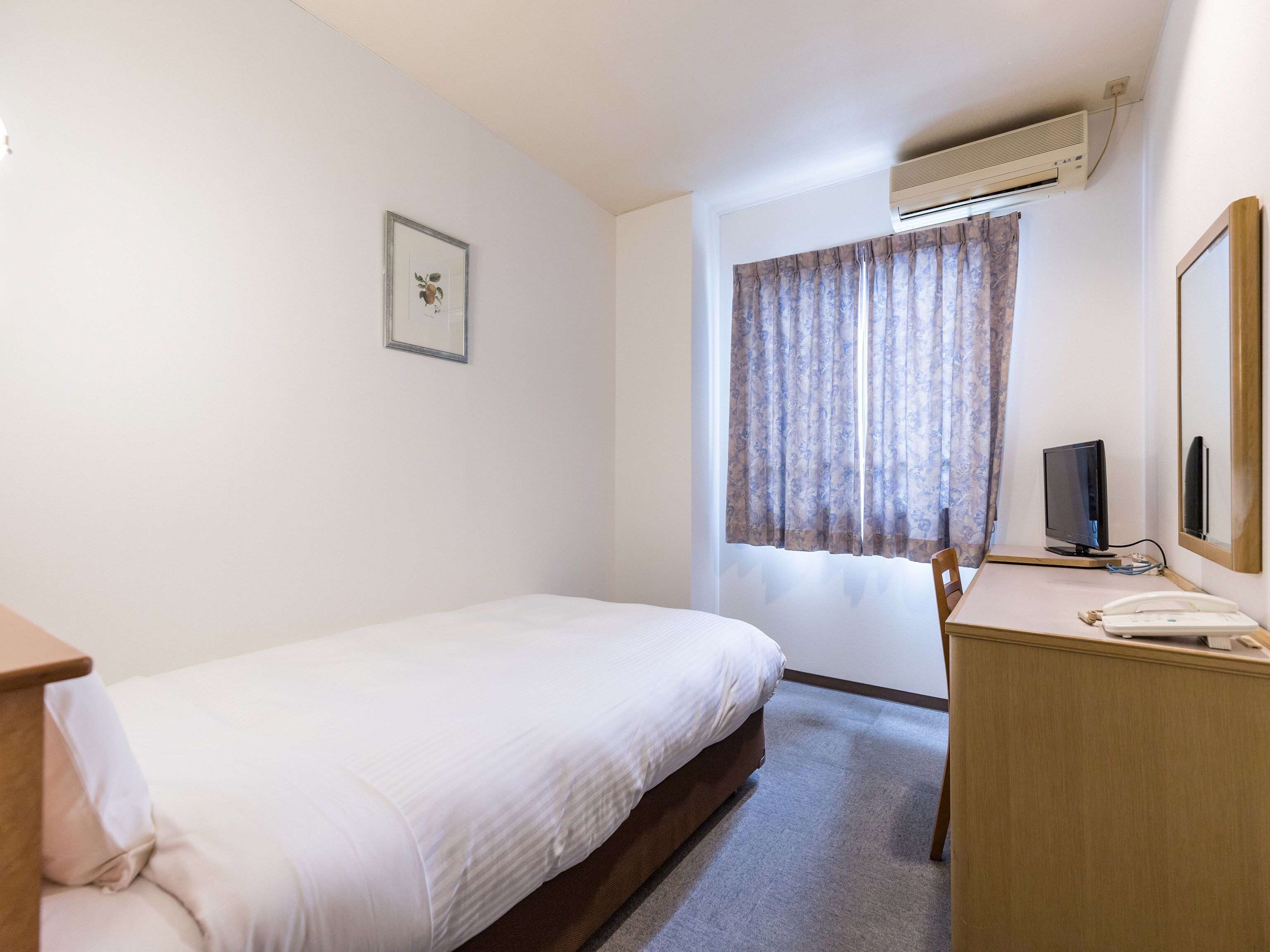 A single bed with a wooden headboard, a desk with a telephone, a television, and a window with curtains.