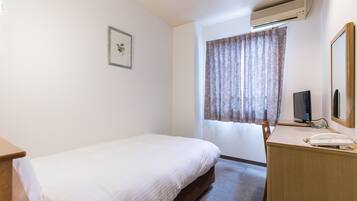 Uma cama de solteiro com cabeceira de madeira, uma escrivaninha com telefone, uma televisão e uma janela com cortinas.