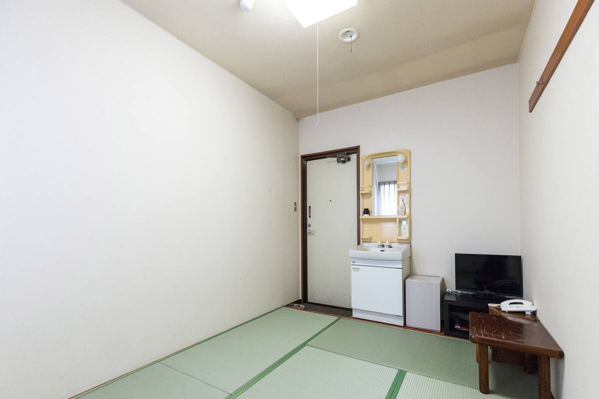 Ein kleiner Raum mit Tatami-Boden, einem weißen Waschbecken, einem Spiegel, einem Fernseher und einem kleinen Holztisch.