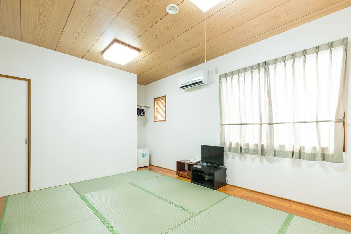 Ein Zimmer mit Tatami-Bodenbelag, Holzbalkendecke, einem fenster mit Vorhängen, einer Wand-Klimaanlage und einem kleinen Regal mit Telefon und Fernbedienung.