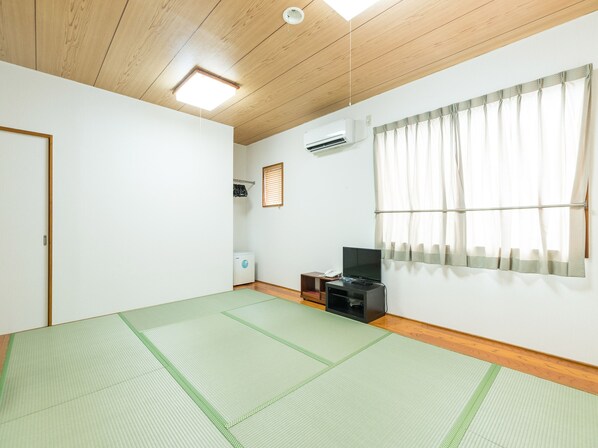 Una habitación con suelo de esteras de tatami, techo de madera, ventana con cortinas, aire acondicionado instalado en la pared y un pequeño estante con un teléfono y un control remoto.