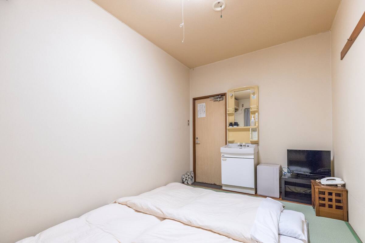 Ein kleines, sauberes Zimmer mit einem Bett, einem Waschbecken, einem Spiegel, einem Fernseher und einem kleinen Tisch.