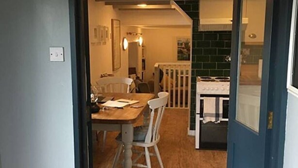 Un comedor con mesa y sillas de madera, barandal blanco y vista hacia una cocina con salpicadero verde.