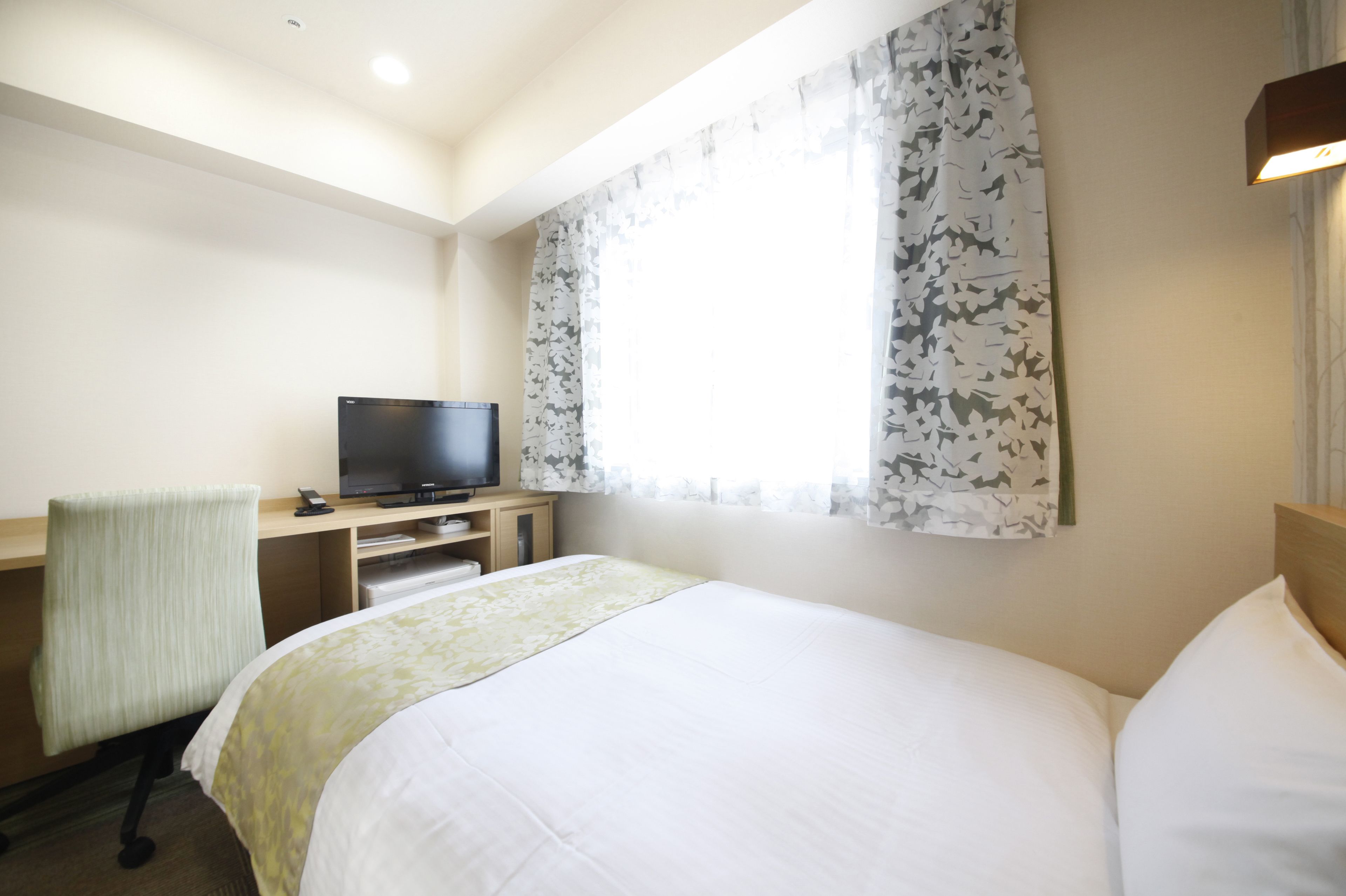 A hotel room with a bed, a desk with a television, and a window with floral curtains.