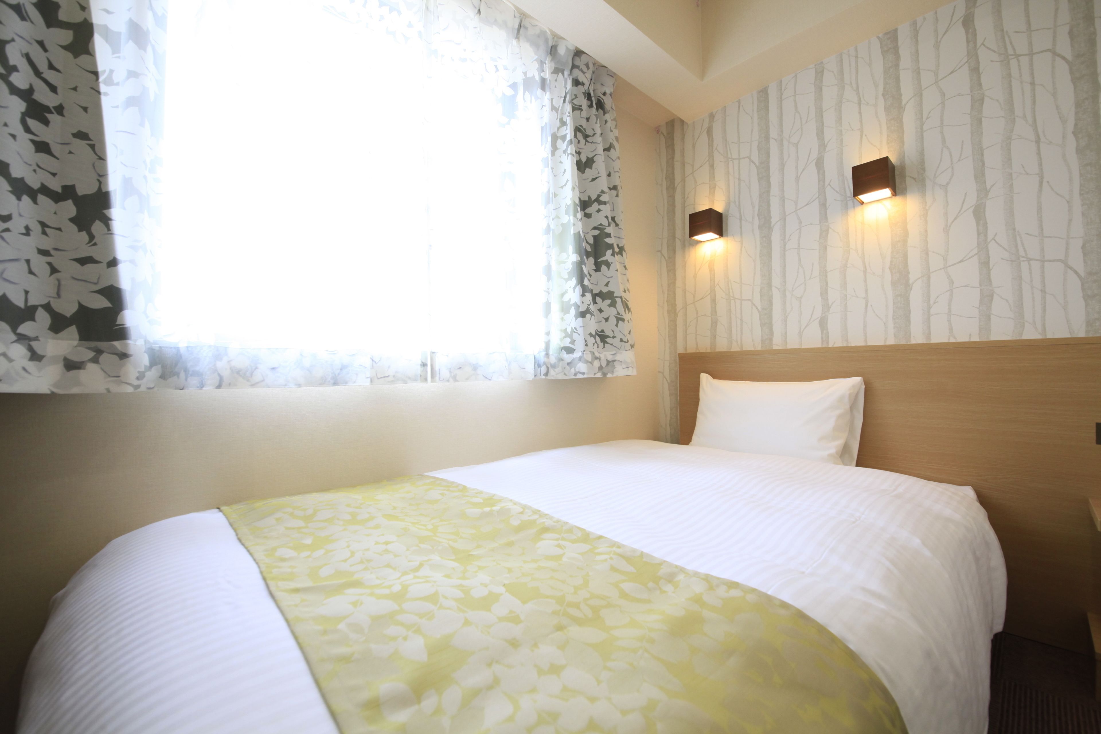 Una habitación de hotel con cama, ventana con cortinas translúcidas y dos luces fijadas a la pared.