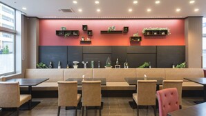 Ein modernes Restaurant mit einer roten Akzentwand, verschiedenen Sitzmöglichkeiten wie Boxen und Tische sowie dekorativen Regalen mit Pflanzen und Flaschen.