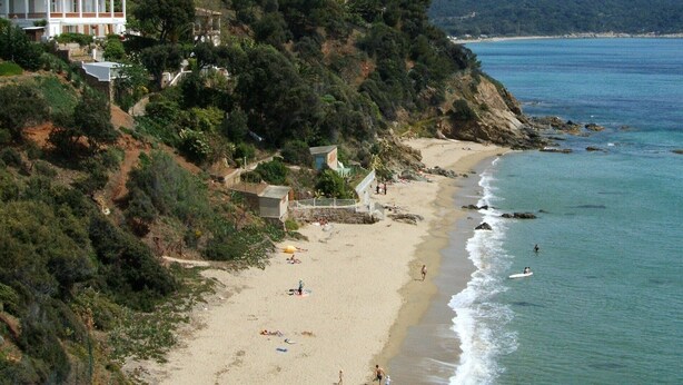 Una playa con un edificio en una colina, una orilla arenosa y el mar.