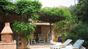 Un patio con horno de ladrillo, sillones blancos y una pérgola cubierta de follaje.