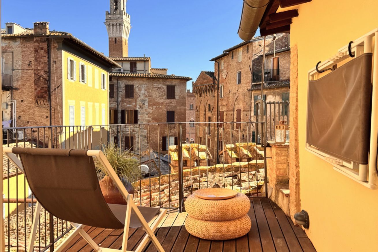 A balcony with a chair and ottoman, overlooking a courtyard with buildings and a tower.