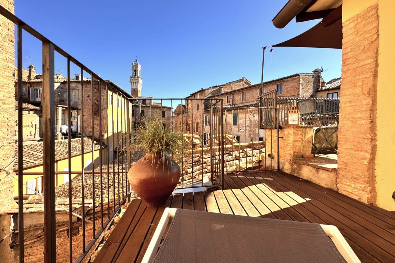 Una terrazza sul tetto con vista su edifici storici e una torre, con un vaso di terracotta grande e una ringhiera in metallo.