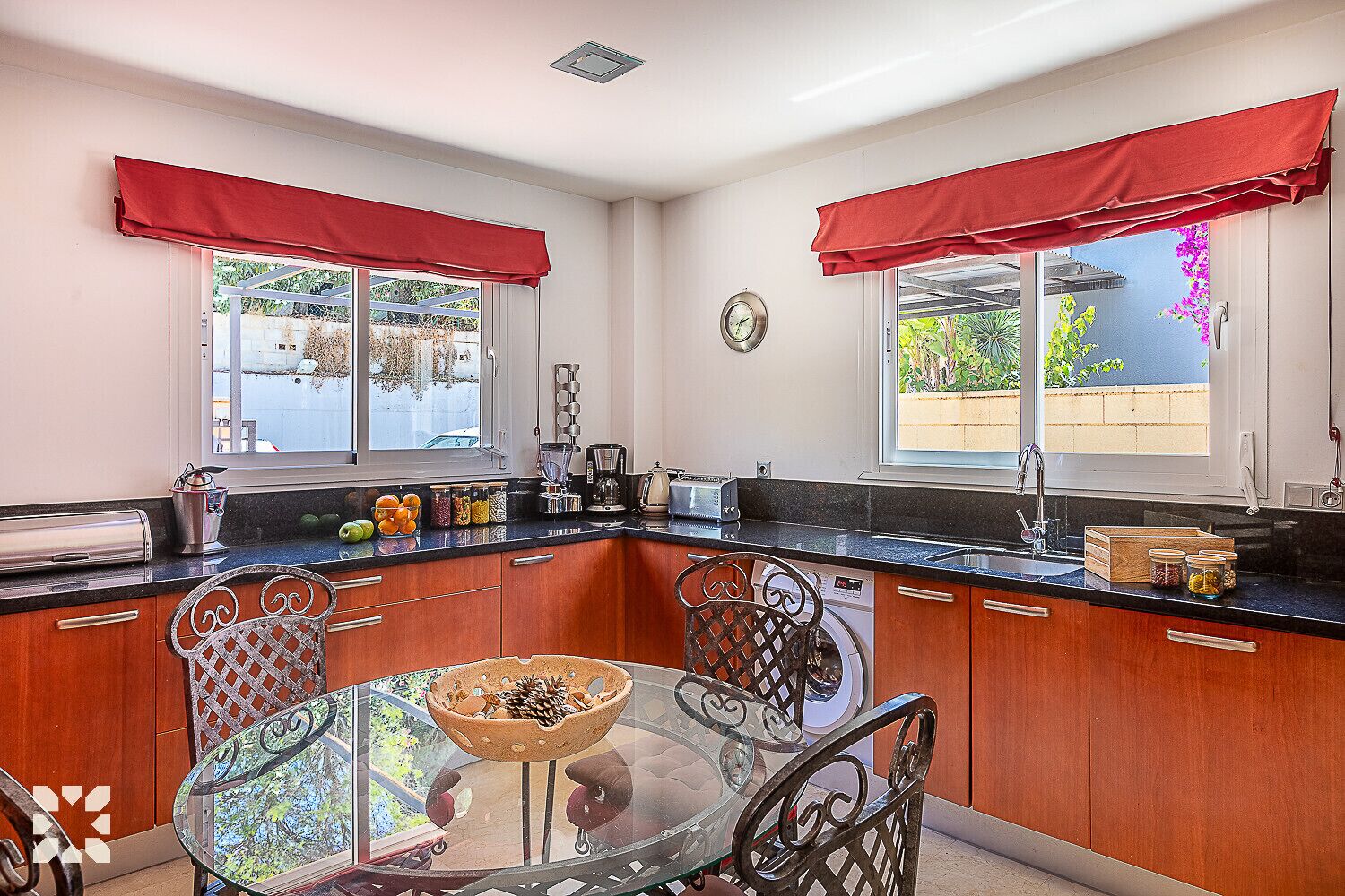 A kitchen with wooden cabinets, a glass dining table, and red window valances.