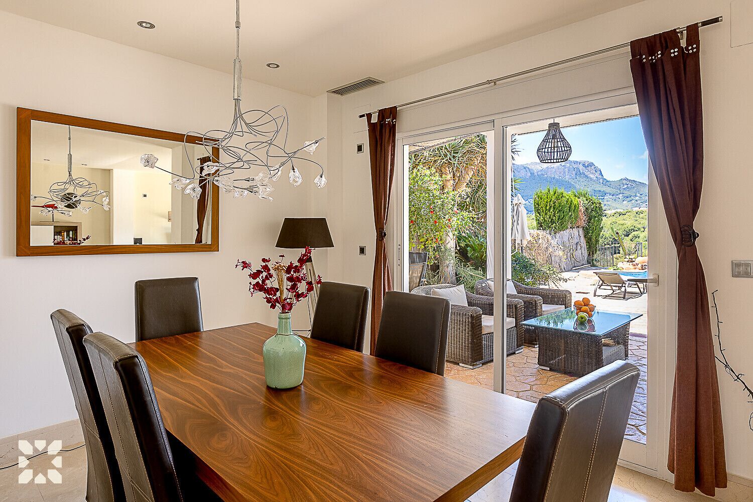 Un coin repas avec une table en bois, des chaises, un vase de fleurs, une lampe et une vue sur une terrasse extérieure aménagée avec des sièges et une piscine.