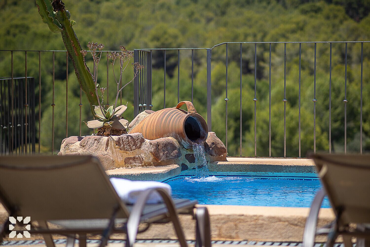 A swimming pool with a water feature, surrounded by a metal fence and overlooking a forested area.
