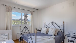 Een slaapkamer met een bed, een raam met gordijnen, uitzicht op een landschap en een kleine tafel met een lamp.