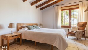 A bedroom with a wooden bed, a nightstand, a chair, and a window with curtains. - Villa SANDRO by Abahana Villas (Benissa)