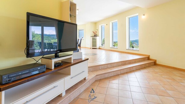 Un salon spacieux avec un téléviseur à écran plat posé sur un meuble en bois, un meuble de rangement blanc et une vue sur l’extérieur grâce à de grandes fenêtres.