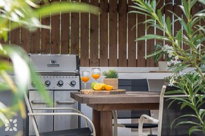 Un espace extérieur aménagé pour les repas, avec un barbecue, une table, des chaises et un pot de fleurs.