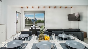 A dining area with a table set for four, a black sofa, a TV mounted on the wall, and a window with a view of mountains and palm trees.