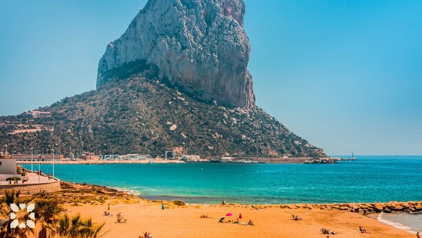 A coastal scene with a prominent rock formation, a sandy beach, and a clear blue sea.