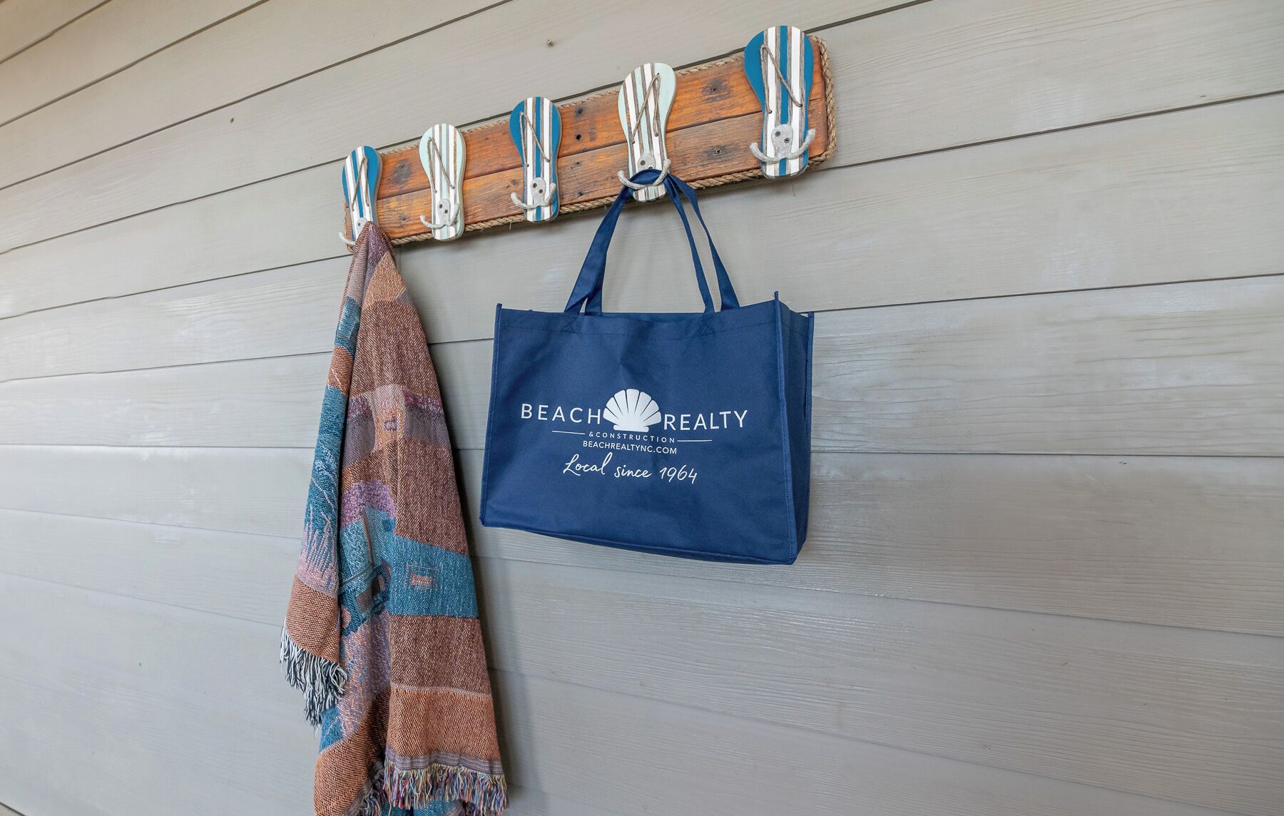 A blue tote bag with "Beach Realty & Construction" printed on it, hanging on a wooden wall with a striped towel and a rope.