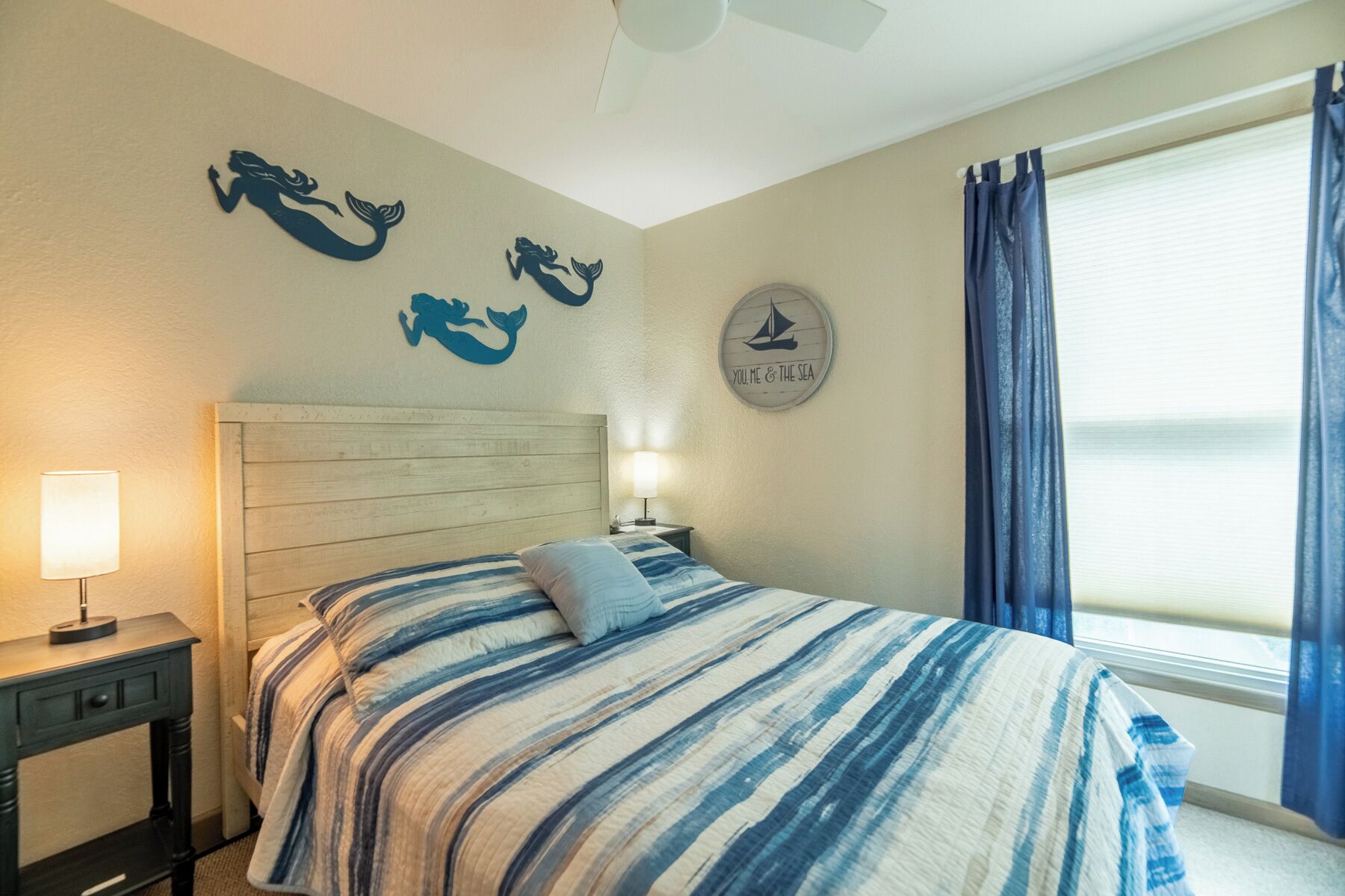 A bedroom with a striped bedspread, a wooden headboard, a nightstand with a lamp, and a wall with mermaid silhouettes and a sailboat sign.