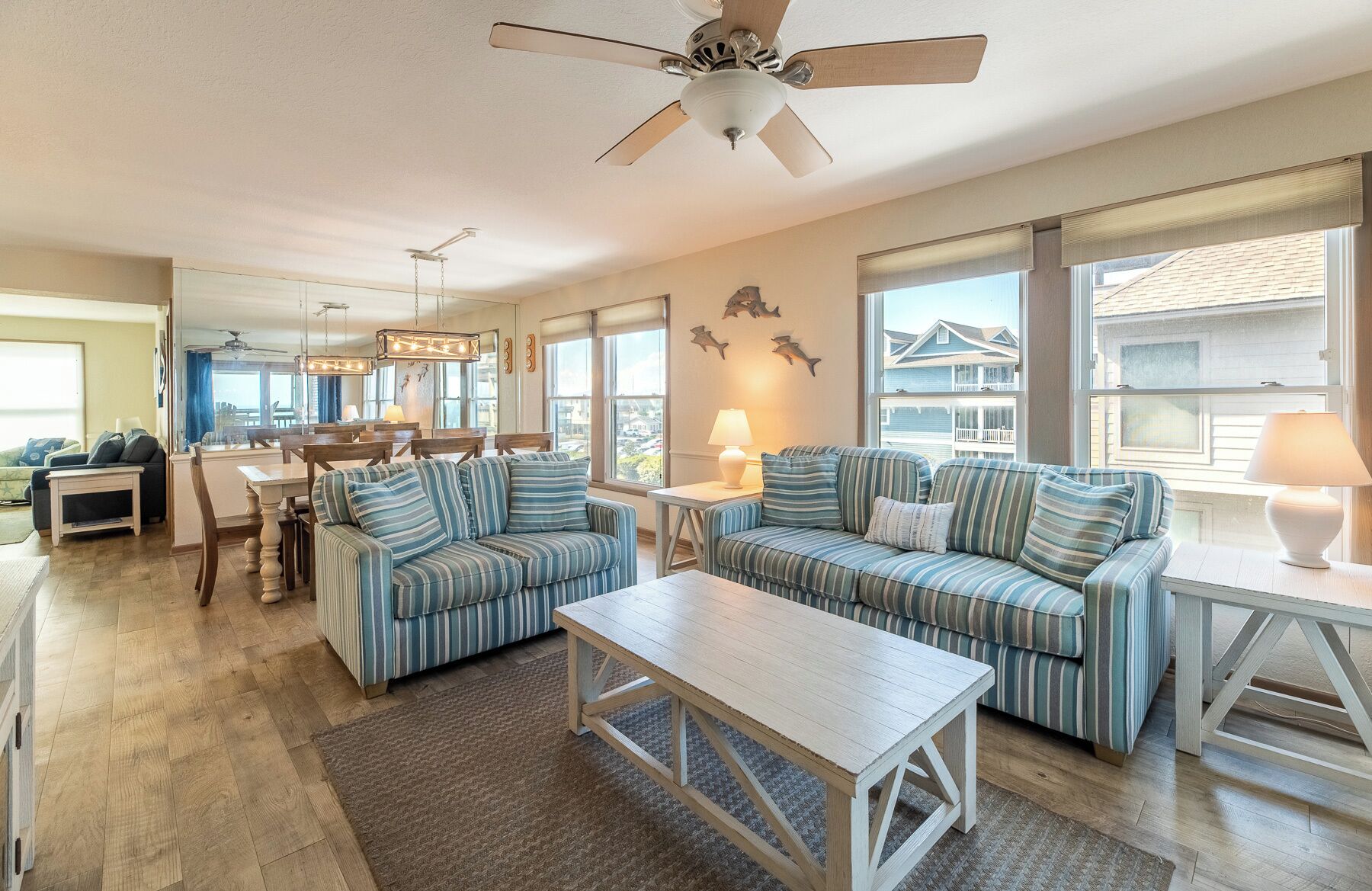 A spacious living room with a ceiling fan, striped sofas, a dining area with a table and chairs, and large windows offering a view of the neighborhood.