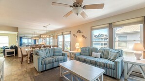 A spacious living room with a ceiling fan, striped sofas, a dining area with a table and chairs, and large windows offering a view of the neighborhood.