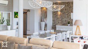A modern living room with a beige sofa, white chairs, a dining table, and a kitchen area with a stone backsplash. - Villa PUNTA VISTA by Abahana Villas (Benitachell)
