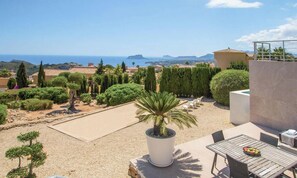 A patio with a table and chairs, a potted plant, and a view of the sea and distant hills.