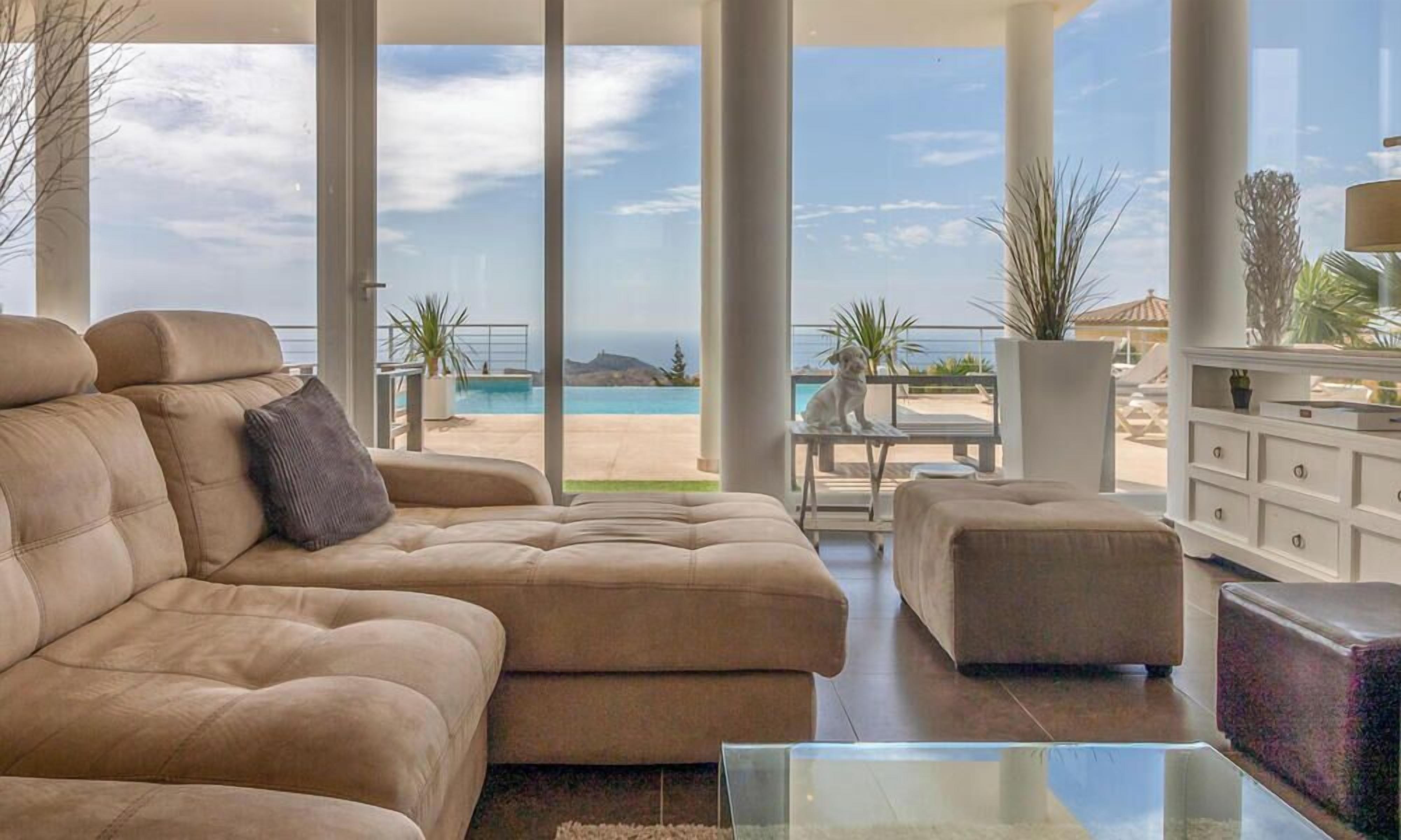 A modern living room with a large beige sectional sofa, a glass coffee table, and a view of a pool and beach.
