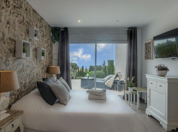 A bedroom with a large bed, a TV, a balcony with a view, and a stone wall. - Villa PUNTA VISTA by Abahana Villas (Benitachell)