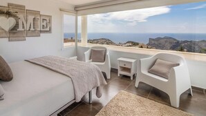 A bedroom with a bed, two chairs, a nightstand, and a view of the sea. - Villa PUNTA VISTA by Abahana Villas (Benitachell)