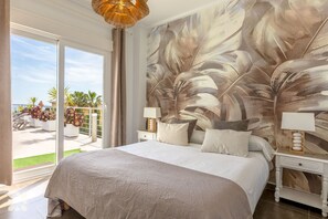 A bedroom with a large bed, two bedside tables with lamps, a view of the outdoors, and a tropical leaf mural on the wall.