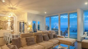 A modern living room with a large beige sofa, a glass coffee table, and a view of the sea through large windows. - Villa PUNTA VISTA by Abahana Villas (Benitachell)